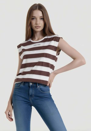 Giovane donna con capelli castani lunghi che indossa un top marrone e bianco a righe senza maniche e jeans blu, in piedi con una mano sui fianchi.