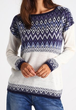 Kvinde iført en hvid strikket sweater med blå og lilla geometriske mønstre på brystet, ærmekanterne og kanten, kombineret med blå jeans.