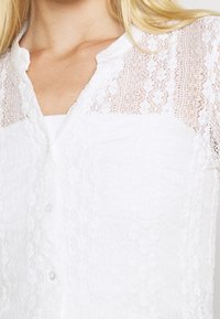 Bílá krajková blůza s knoflíkovým zapínáním, jemným květinovým vzorem a průhlednou krajkovou částí na ramenou. Měkká textura s hladkým povrchem.