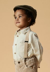 Beige overhemd met knopen, beige corduroy broek met bretels, groene pet, krullend haar. Eenvoudig ontwerp met een detail van een borstzak.