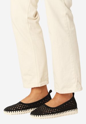 Voeten dragen zwarte mesh instapschoenen met witte, getextureerde zolen, gecombineerd met off-white rechte pijpen broeken.