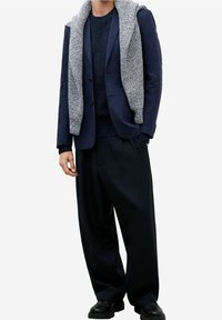 Blazer navy superpuesto a un suéter oscuro y texturizado, combinado con pantalones negros holgados. Una sudadera de punto gris está colgada sobre el blazer. Zapatos negros.