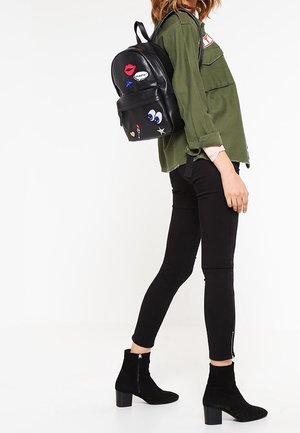 Vrouw die zwarte strakke broek, zwarte laarzen met hak, groene jas en zwarte rugzak met kleurrijke patches draagt, staand in profiel.