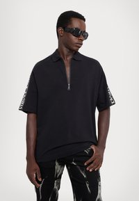 Čierne polo tričko s krátkym rukávom a štvrtinovým zipsom, s detailom loga na rukávoch, v kombinácii s čiernymi nohavicami so bielym abstraktným vzorom.