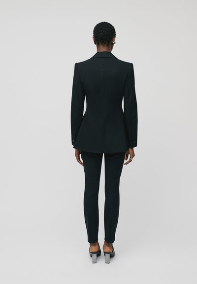 Črn suknjič z oblikovanimi rameni in ukrivljenim robom, v kombinaciji z ozkimi hlačami. Material deluje gladko, kar poudarjajo elegantni čeveljci.