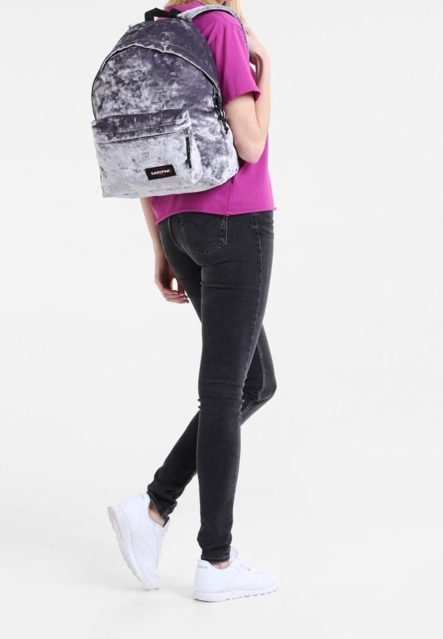 Personne portant une chemise violette, un jean skinny noir, des baskets blanches et un sac à dos Eastpak en velours gris dans le dos.