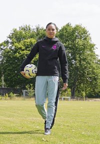 Melna jaka ar purpursarkanām akcentiem, gaiši pelēkas bikses ar melnām sānu svītrām, tur rokās futbola bumbu, stāv zālājā ar kokiem fonā.