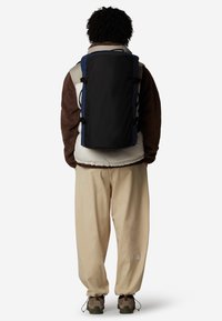 Vista posteriore di uno zaino nero con dettagli blu, abbinato a un fleece marrone e un giubbotto trapuntato crema, pantaloni beige e sneaker grigie.