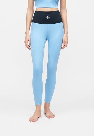 Γυναίκα φοράει ανοιχτό μπλε κολάν με ψηλή μέση και μαύρη μέση με λευκό λογότυπο Calvin Klein, στέκεται ξυπόλητη σε απλό φόντο.