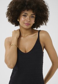 Melnas adītas bez piedurknēm topiņš ar dziļu V-veida izgriezumu un šaurām siksniņām. Audumam ir struktūra ar horizontālu rakstu. Akcentēts ar zelta aproci.