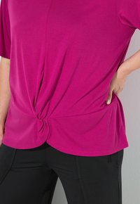 Top fucsia de manga corta con un detalle anudado en la cintura, combinado con pantalones negros con bolsillos y un diseño ceñido.