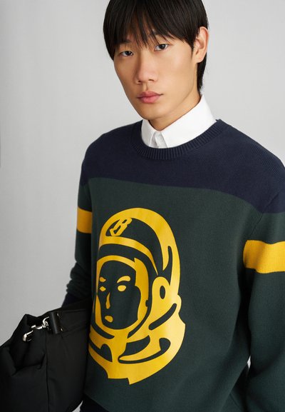 Suéter de punto con diseño de bloques de color en navy y verde, gráfico de astronauta amarillo y cuello redondo acanalado. El modelo sostiene un bolso negro.
