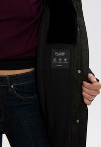 Sort polstret jakke med en struktureret krave, der har et mærke, som angiver vandmodstand, vindmodstand og termisk isolering.