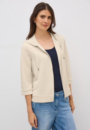 Light beige zip-up hoodie with a hood, three-quarter sleeves, and drawstrings, worn over a navy tank top. Soft fabric with a smooth texture.