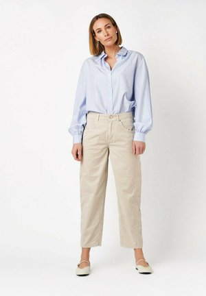 Woman standing wearing light blue striped blouse with puffed sleeves, beige high-waist pants, and cream flat shoes against white background.