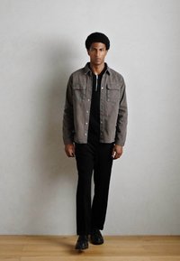 Chaqueta gris de botones sobre una camiseta negra, combinada con pantalones y zapatos negros. La chaqueta cuenta con dos bolsillos frontales y una textura suave.