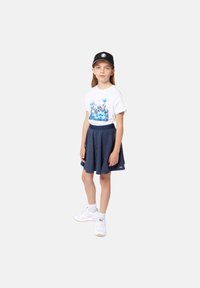 T-shirt blanc avec un graphisme coloré, jupe plissée bleu marine, baskets blanches et casquette noire. Texture en coton lisse avec une coupe décontractée.