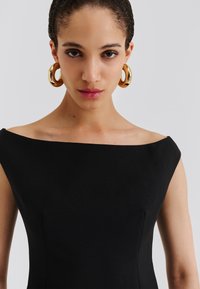Čierne šaty s odhalenými ramenami, lichotivým strihom a hladkou textúrou. Celý vzhľad dopĺňajú zlaté kruhové náušnice.