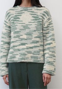 Maglione lavorato a maglia color crema con motivo camouflage verde, dotato di scollatura a girocollo e polsi a coste. Indossato con pantaloni verdi abbinati.