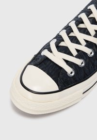 Platnene tenisice s teksturiranim tamnoplavim gornjim dijelom, kremasti gumeni zaštitni vrh i bijele vezice. Ima metalne prolaze i crni rub duž potplata.