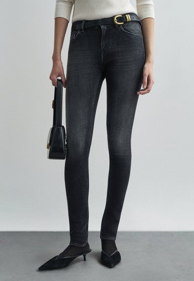 Jeans skinny in denim nero con una leggera scoloritura, dotati di cinque tasche, passanti per cintura e una fibbia dorata, abbinati a tacchi a punta neri e a una piccola borsa.