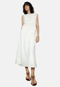 Robe maxi blanche en lin avec un corsage en dentelle texturée, design sans manches et coupe décontractée. Associée à des sandales brunes.
