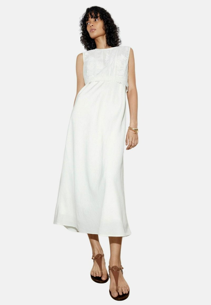 Robe maxi blanche en lin avec un corsage en dentelle texturée, design sans manches et coupe décontractée. Associée à des sandales brunes.