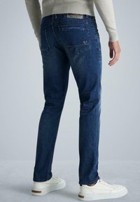 Vaqueros de mezclilla azul de corte ajustado con un acabado ligeramente desteñido, cinco bolsillos y una etiqueta en la cintura; combinados con zapatillas blancas.