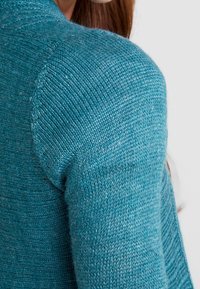 Stickad tröja i teal, med en närbild av det strukturerade tyget och axelsömmen, som har detaljer av fint garn och ribbade accenter.