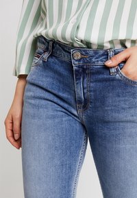 En person som bär en grön och vit randig blus poserar med en hand på midjebandet av blå denimjeans.