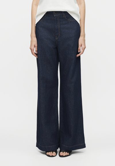 Personne portant un jean large bleu foncé, un haut blanc et des sandales noires à bouts ouverts, debout devant un fond clair uni.