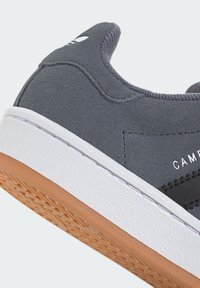 Zapatilla de ante gris con suela de goma blanca y marrón. Presenta un talón redondeado, parte superior texturizada y costuras en contraste. Marca visible en el lateral.