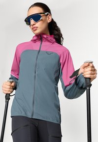 Veste légère gris et rose, avec une fermeture éclair à l'avant, une capuche et des trous de ventilation. Détail de logo noir sur la poitrine. Bâtons de trekking à la main.