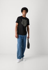 Camiseta negra de manga corta con un gráfico de león blanco, combinada con jeans de mezclilla azules y zapatillas blancas; sostiene una bolsa negra.