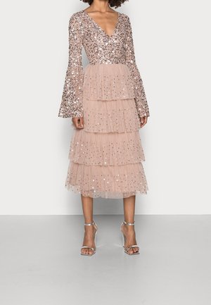 Woman wearing a long-sleeve, sequined pink dress with tiered tulle skirt and silver high-heeled sandals, standing against a plain background.