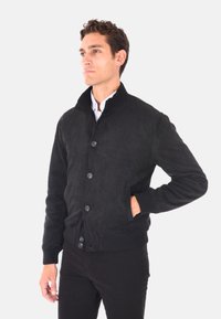 Giacca bomber in suede nera con colletto rialzato, polsini a costine, chiusura frontale con bottoni e tasche laterali. Tessuto morbido e design aderente.