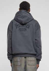 Grijze oversized hoodie met een capuchon, voorzien van een opvallende zwarte grafische tekst op de achterkant. Gemaakt van zachte stof met een gladde textuur.