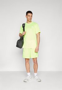 Limegrünes Sport-T-Shirt und Shorts, aus leichtem Stoff, Rundhalsausschnitt, Logo auf der Brust, kombiniert mit einer schwarzen Tasche über einer Schulter.