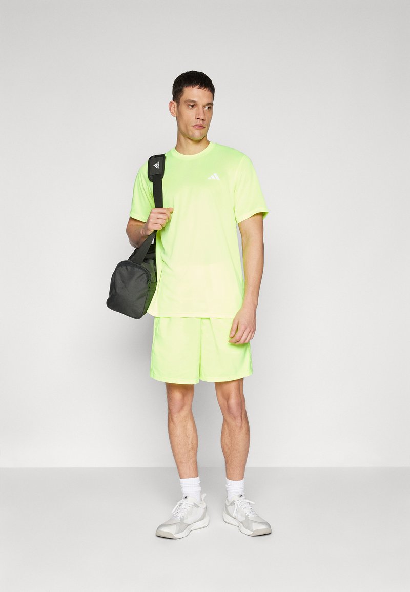 T-shirt e pantaloni sportivi di colore verde lime, in tessuto leggero, colletto rotondo, logo sul petto, abbinati a una borsa nera a tracolla.