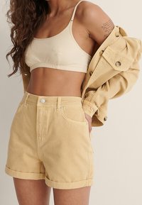 Shorts de denim amarillos con dobladillos enrollados, bolsillos frontales y cierre con botón de metal, combinados con una blusa corta de color beige claro y una chaqueta a juego.