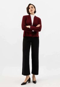 Cardigan bordeaux con finitura testurizzata, indossato sopra una maglietta bianca, abbinato a pantaloni neri a gamba larga e scarpe con tacco nere. Design semplice e moderno.