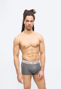 Homme torse nu aux dreadlocks longs portant un boxer gris BOSS, debout devant un fond clair uni, faisant face à l'avant.