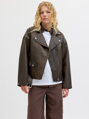 Bruine leren motorjas met grote revers, diagonale rits, voorkwasten en gespsluiting. Gekleed over een wit shirt en gestreepte broek.
