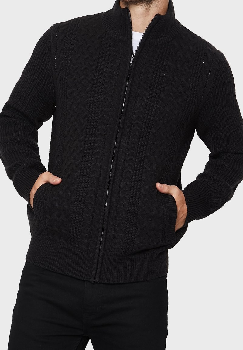 Cardigan noir en tricot avec un col montant, présentant un motif en torsades texturé, une fermeture éclair à l'avant et des poches latérales. Les bords-côtes complètent le design.