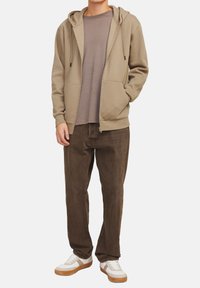 Beige zip-up hoodie med snøretræk i hætten, båret over en taupe t-shirt, parret med mørkebrune bukser og hvide sneakers med brune detaljer.