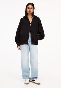 Sweat à capuche noir avec une coupe ample, poignets côtelés, deux poches latérales, associé à un jean large bleu clair et des baskets blanches.