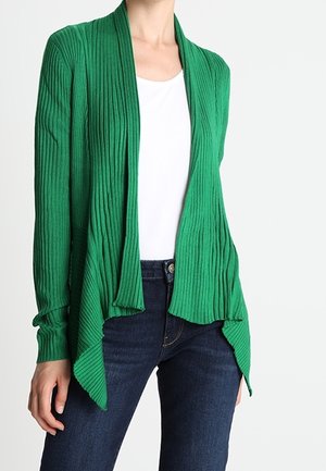 Frau trägt eine grüne, gerippte, offene Strickjacke über einem weißen Oberteil und dunkelblauen Jeans, steht vor einem schlichten hellen Hintergrund.