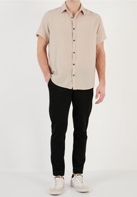 Camisa beige de manga corta con cierre de botones negros en la parte delantera, combinada con pantalones negros de corte ajustado y zapatillas blancas con detalles beige.
