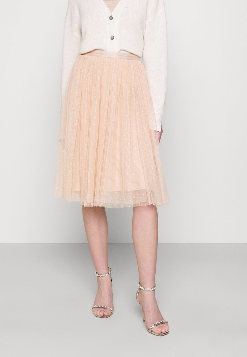 Jupe en tulle pêche avec une silhouette pleine et à étages, ornée de motifs en pois blancs. Portée avec un cardigan crème et des sandales à talons argentés.