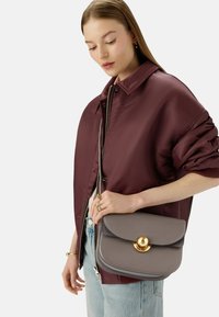 Borsa a tracolla in pelle grigia con alette arrotondate e dettagli in metallo dorato, tenuta da una persona con un bomber bordeaux e jeans blu.